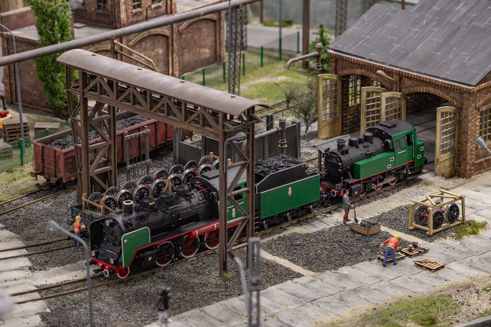 A miniature railway scene featuring two steam locomotives, one with exposed wheels under a shed, and the other near a brick engine house, with small figures of workers tending to the coal and tracks.