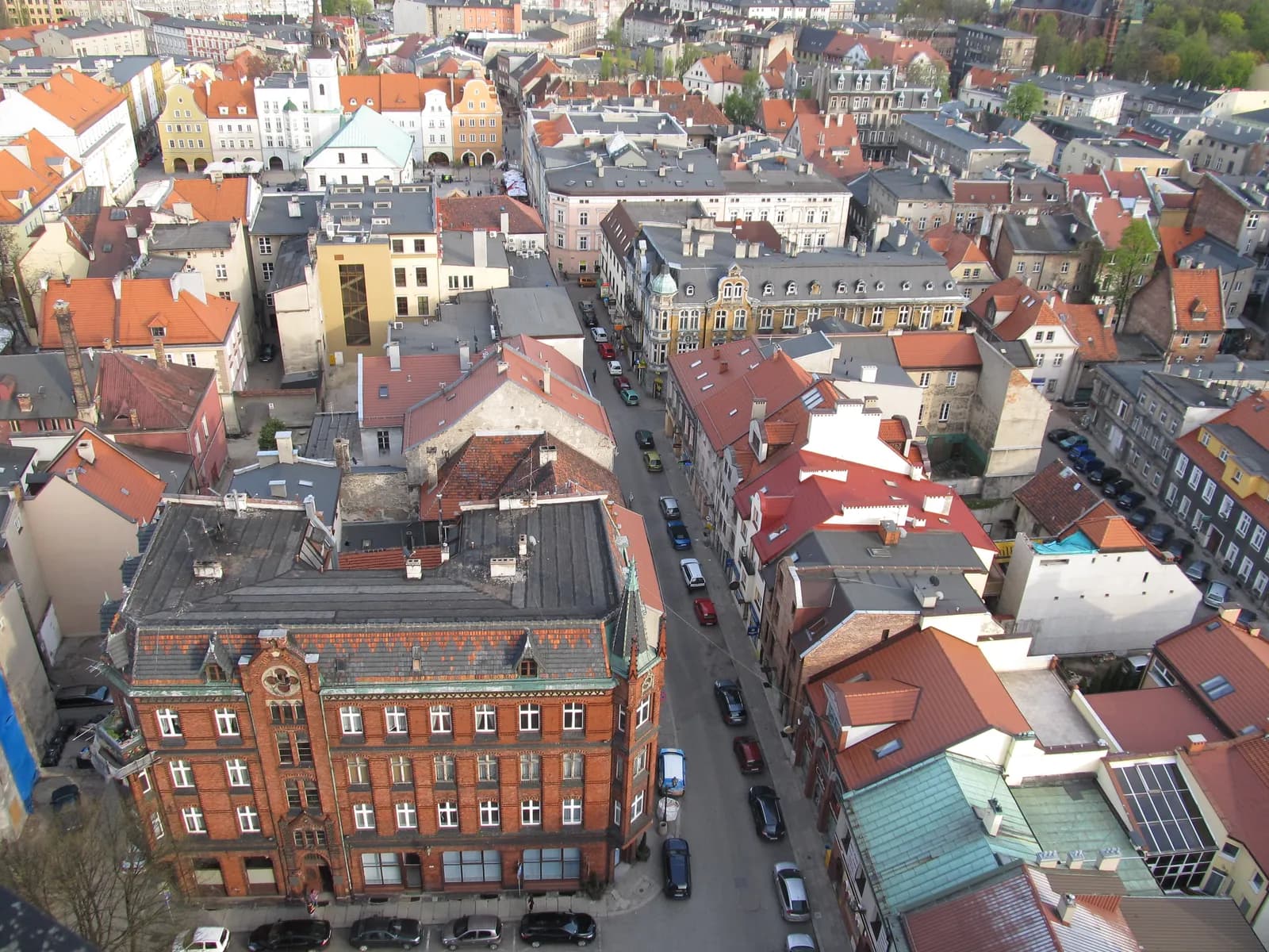 An aerial view of a densely packed historic European city with a mix of red-tiled and dark-roofed buildings, a prominent red brick building in the foreground, and streets with parked cars winding through the urban landscape.
